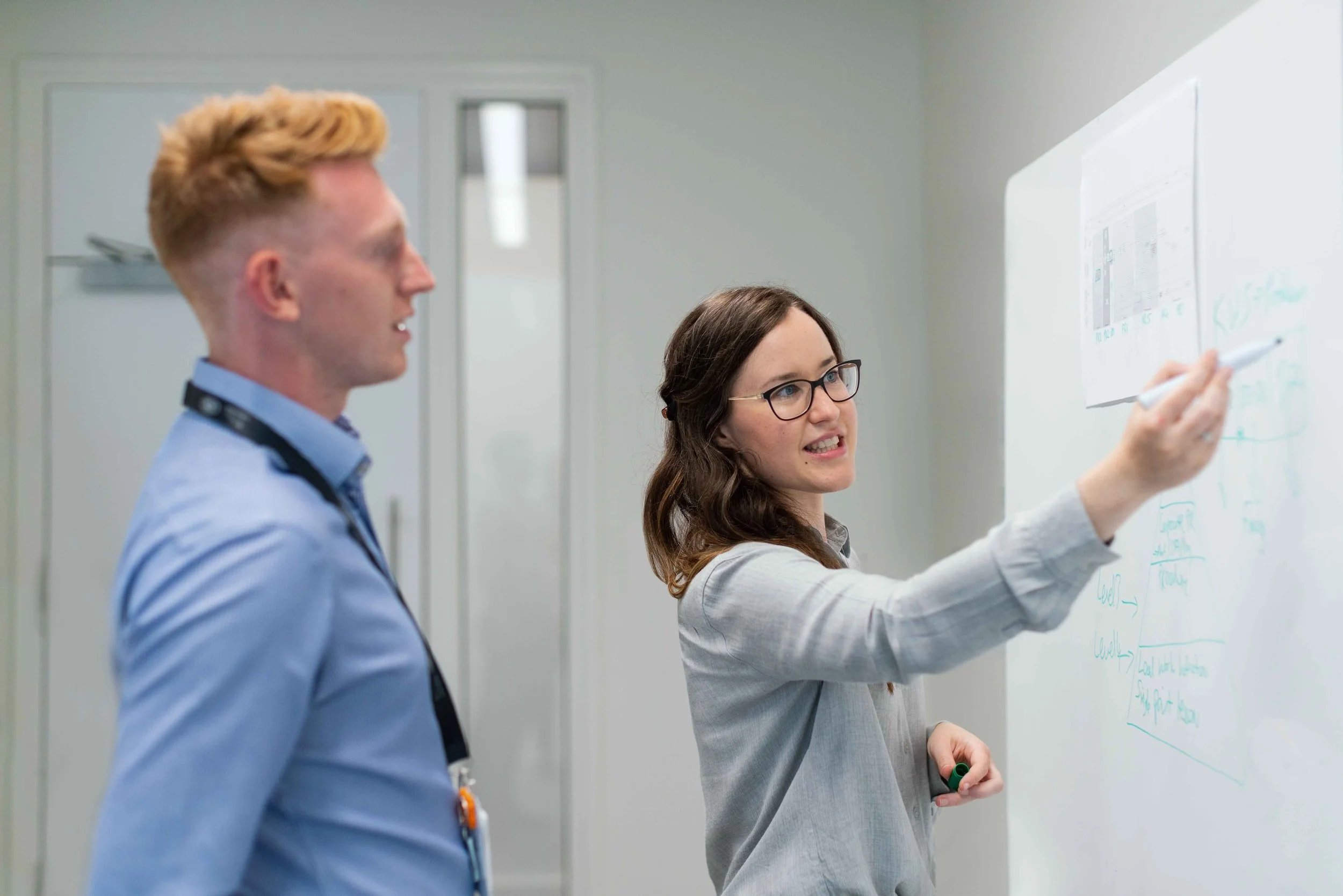 A woman with glasses and brown hair writing on a whiteboard while a man with blond hair and a blue shirt observes.