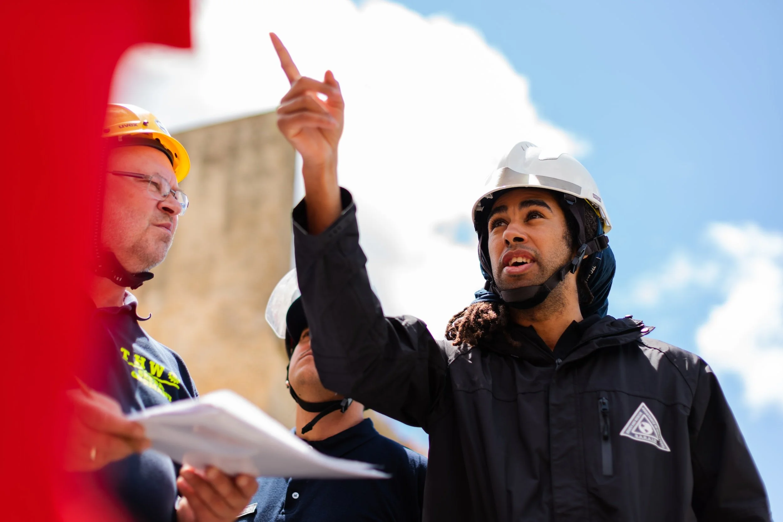 Two construction workers, one older man with glasses and a yellow helmet and a younger man with a white helmet and dreadlocks, are engaged in a discussion outdoors, with a person in the background holding a clipboard.